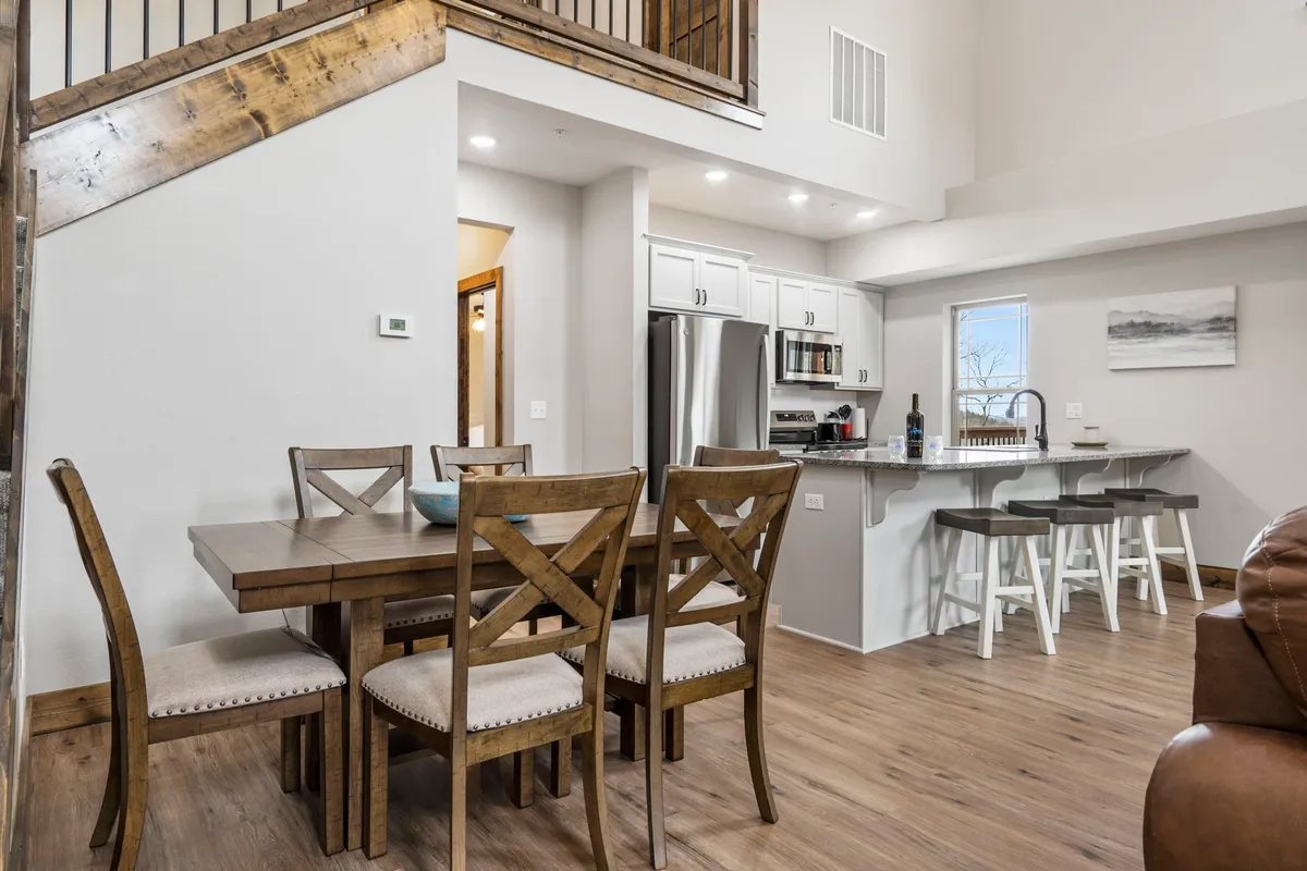 A bright and inviting dining area centered around a large wooden dining table, with a kitchen island and staircase visible in the background. Expansive windows let in abundant natural light, while the layout supports seamless movement between dining, cooking, and relaxing. The setting is ideal for shared meals, celebrations, and conversations that bring families together.