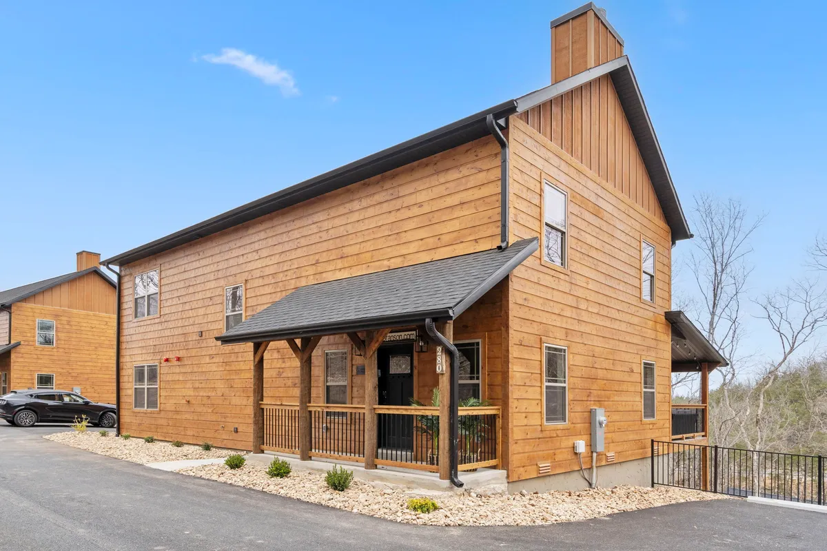 A cozy wooden cabin with a covered porch overlooks a wide driveway and the surrounding natural landscape. The outdoor space is inviting, with evening sunlight suggesting warm sunsets and opportunities for family gatherings.