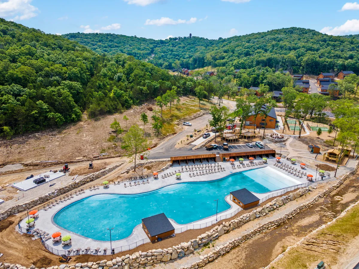 An exterior view of a spacious outdoor swimming pool surrounded by lush greenery, poolside loungers, and cabana structures. The setting is ideal for swimming, sunbathing, and outdoor games, presenting a lively yet serene resort atmosphere.