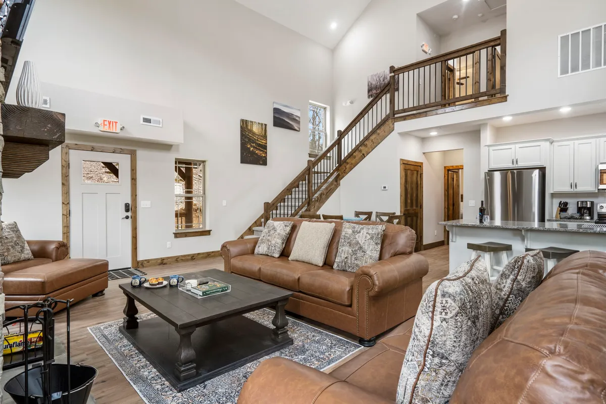 A warm, open-plan living area featuring plush leather sofas arranged around a coffee table, with a clear view of the adjacent kitchen area and a staircase leading to the next level. The overall setup invites group gatherings and family conversations, with the kitchen in easy reach for snacks or drinks. The natural light and cozy furnishings create an atmosphere perfect for relaxing evenings or celebrations.