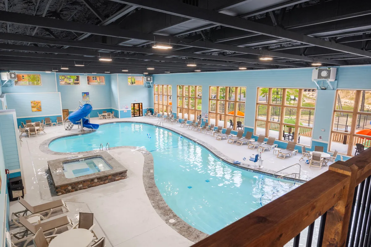 A bright indoor swimming pool area, featuring a sleek water slide, spacious spa section, and large windows letting in natural light. The poolside seating provides space for families to gather and relax, making it an ideal year-round amenity regardless of the weather outside.