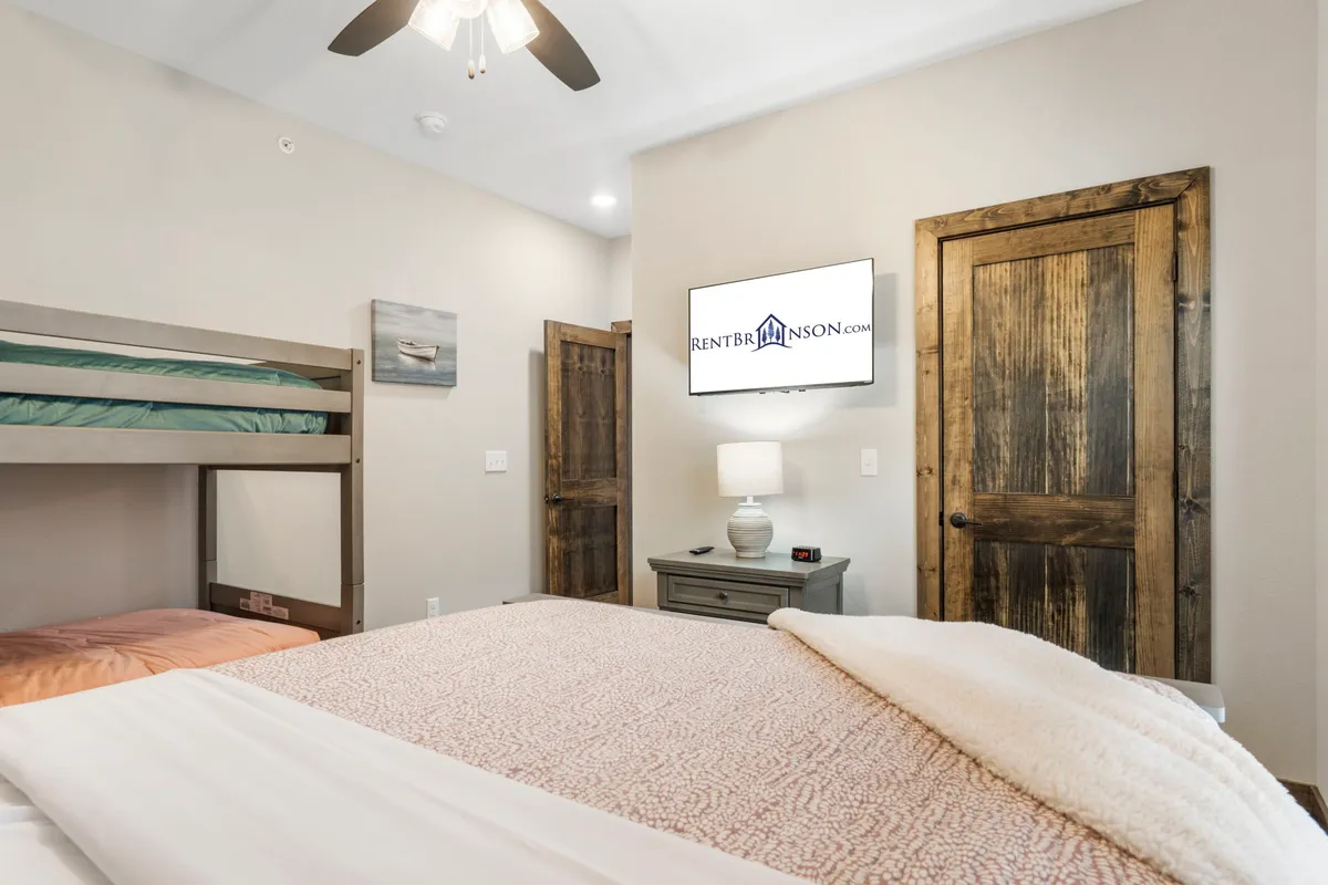 A welcoming, family-oriented bedroom featuring a queen bed and a twin-over-twin bunk bed. The room is bright with a TV, wooden furniture, a nightstand with a lamp, and a cozy, inviting atmosphere ideal for both adults and kids.