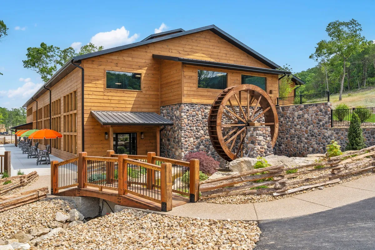 An exterior view of a charming building featuring a wooden water wheel, stone facade, large windows, and an outdoor seating area. The scene suggests opportunities for dining, social gatherings, and peaceful nature walks beside the picturesque feature.