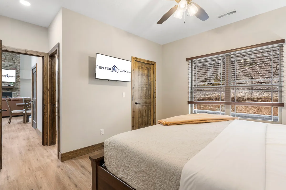 A cozy bedroom featuring a bed with a quilted cover, a television mounted on the wall, large windows with blinds, and a wooden door. The space exudes comfort and modern convenience, ideal for personal retreat after a day of adventure.