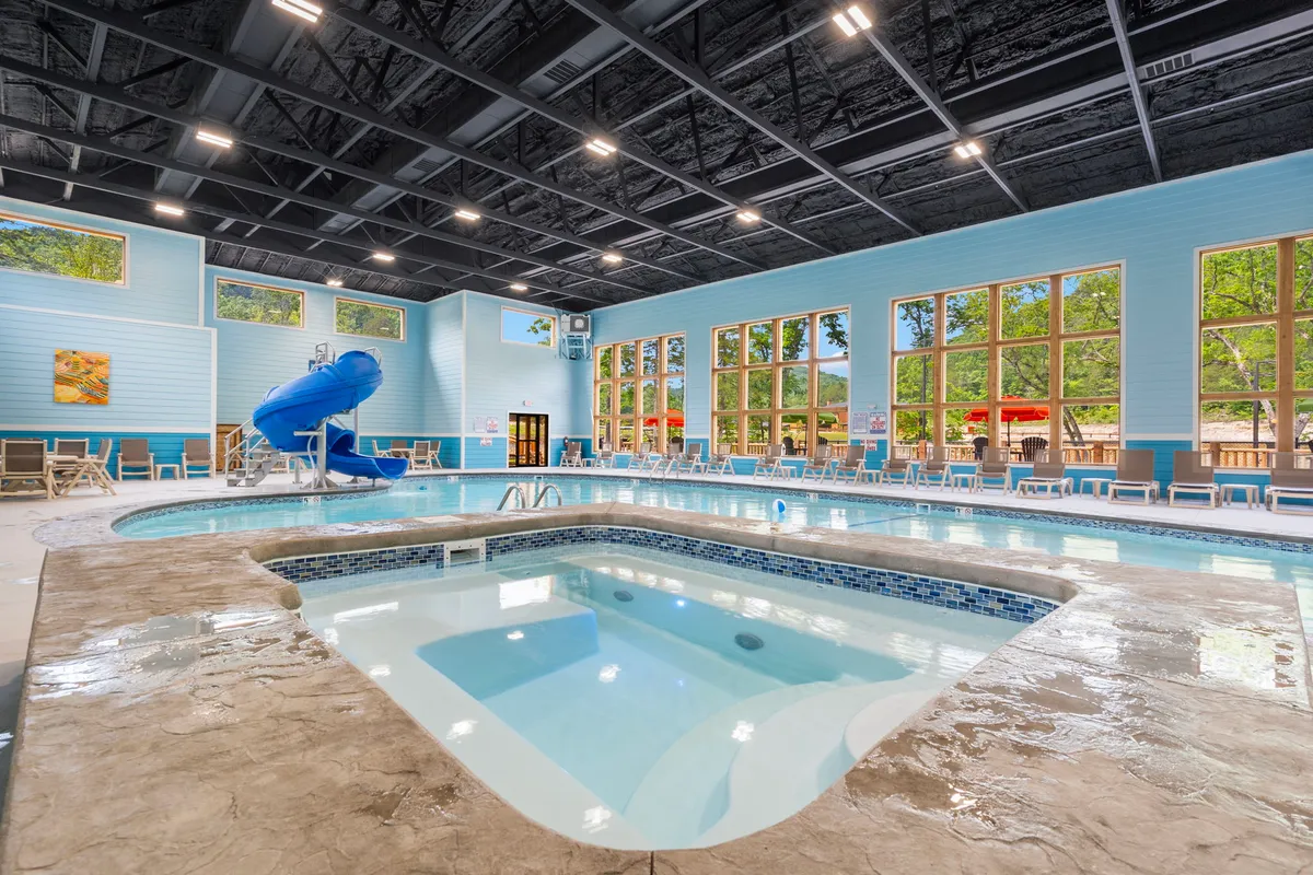 This interior image features a bright indoor pool area complete with a water slide, hot tub, large windows that let in natural light, and poolside lounge chairs. Children play on the slide while others relax in the hot tub, capturing the vibrant atmosphere of year-round aquatic fun.