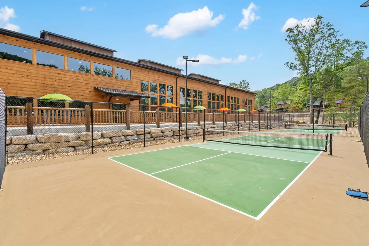 A bright, welcoming exterior view of modern tennis courts bordered by greenery, adjacent to a stylish wooden building with outdoor seating and green umbrellas. The scene highlights opportunities for active recreation and socializing.